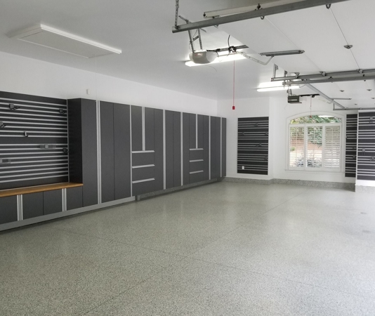 Garage interior featuring charcoal gray and silver cabinets, black-and-white slatwall on white walls, and a light gray speckled epoxy floor.
