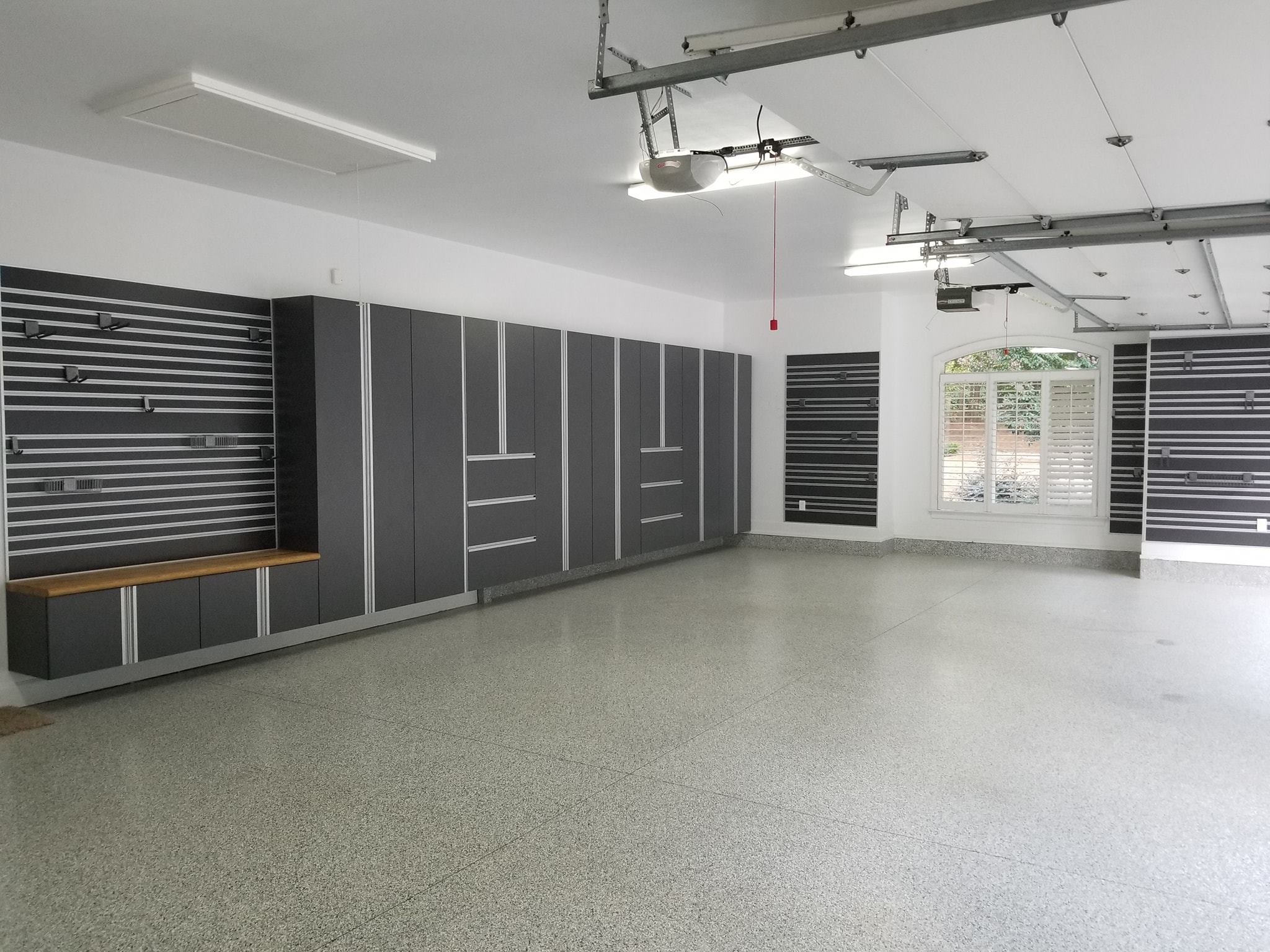 Garage interior featuring charcoal gray and silver cabinets, black-and-white slatwall on white walls, and a light gray speckled epoxy floor.