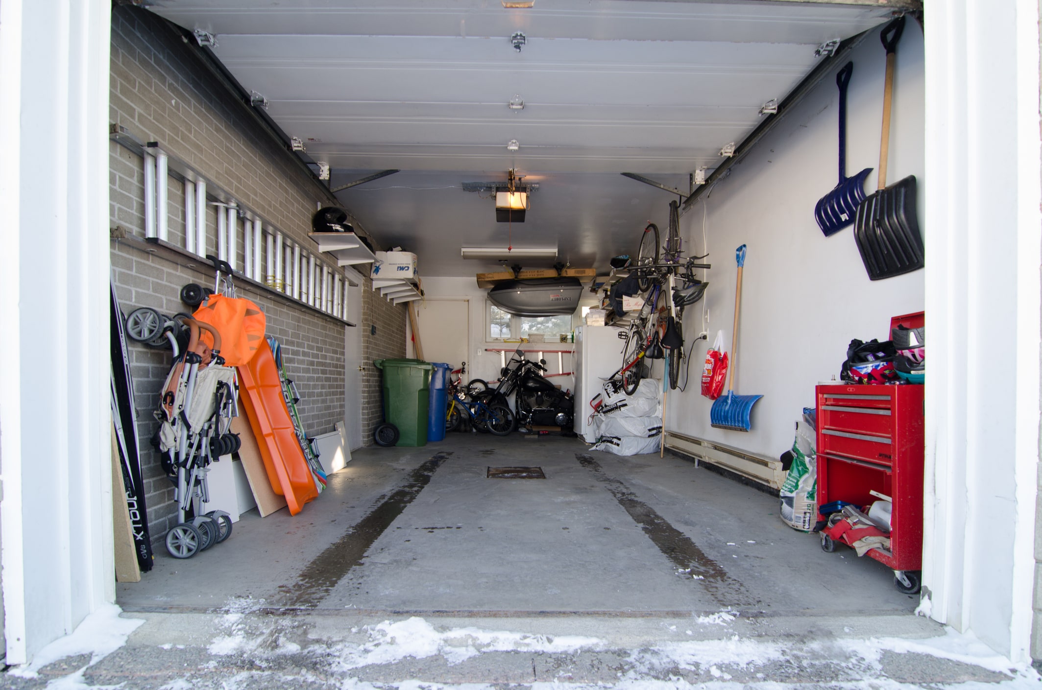 Interior of a residential garage in winter with snow at the entrance, concrete flooring, and storage items. 